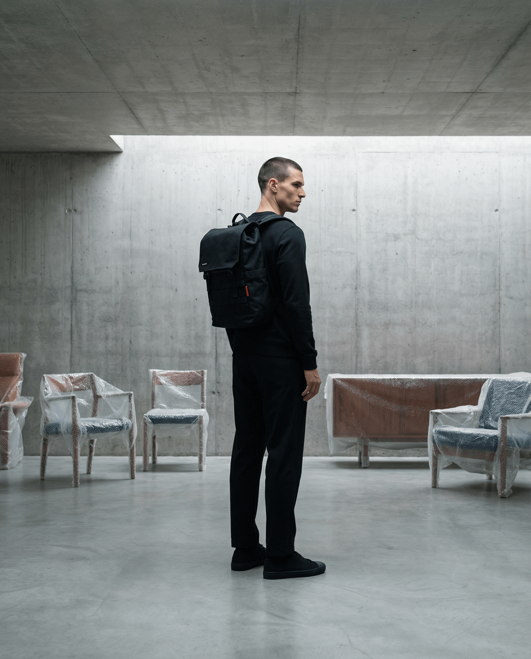 Man wearing a black outfit and Minato Lite 2.0 - Dark Black backpack standing in a modern, minimalist space.