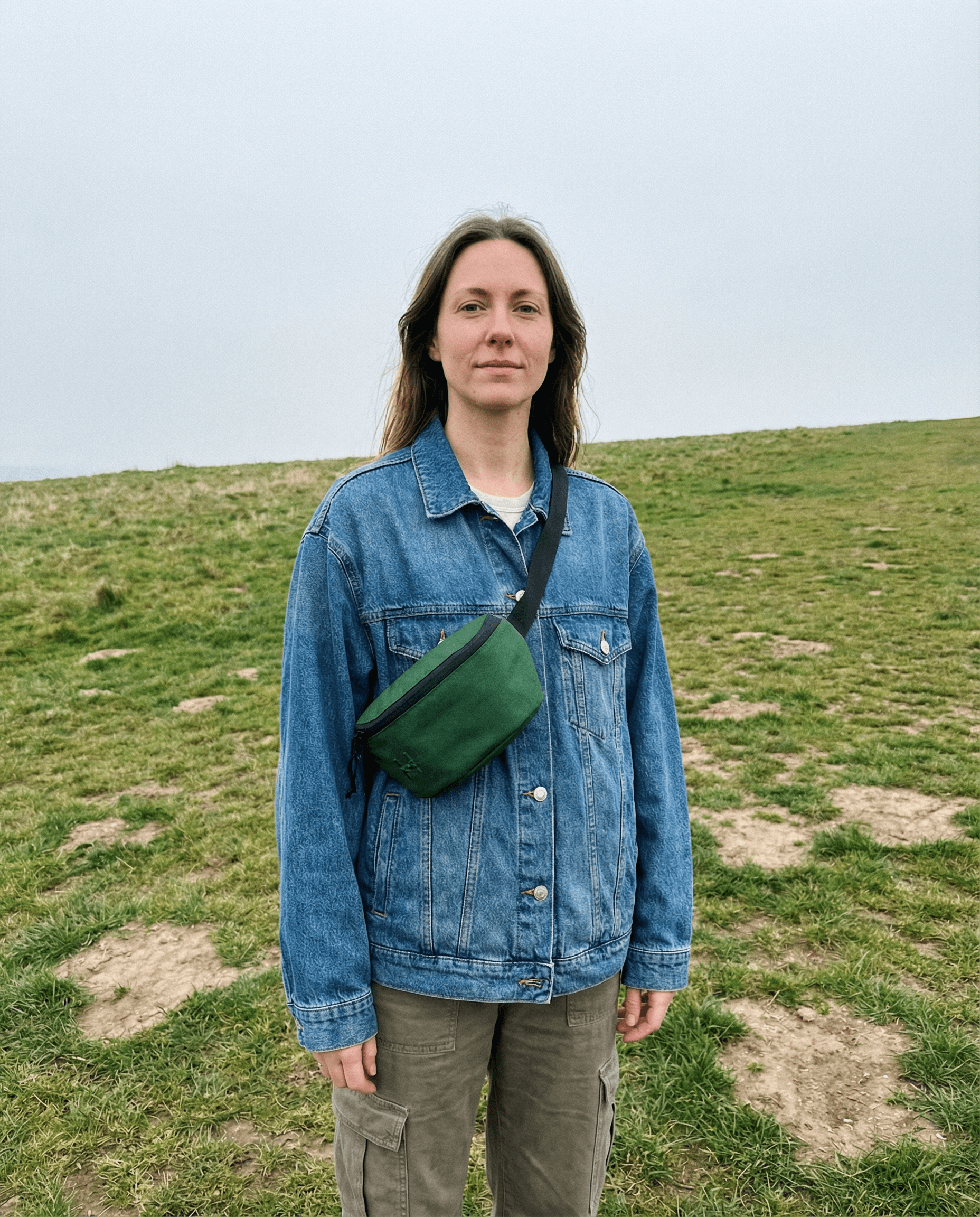 A woman wearing a denim jacket and a Sora 2.0 - Forest Green bag stands on grassy terrain.